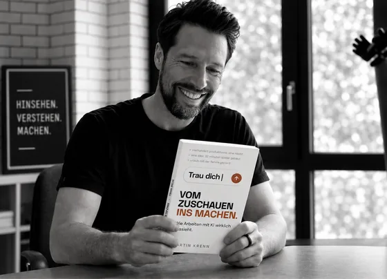 Martin Krenn am Schreibtisch mit dem gedruckten Buch, im Hintergrund ein Plakat mit den Worten Hinsehen, Verstehen, Machen.